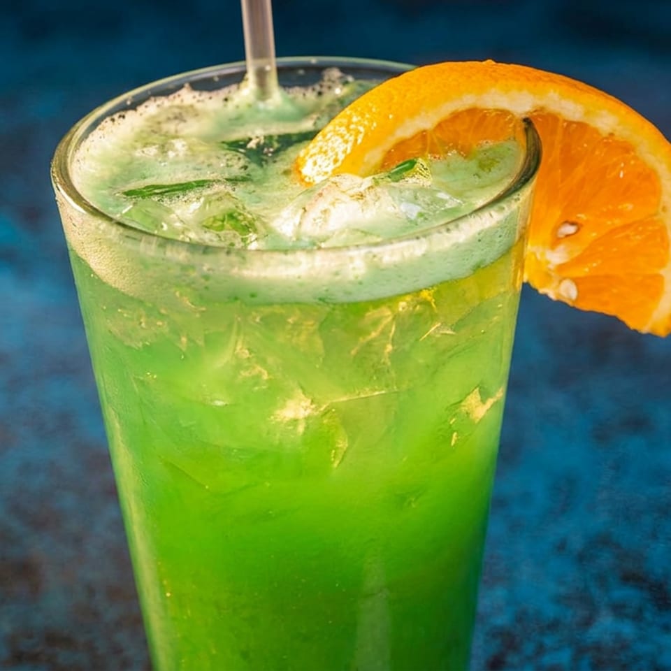 Vibrant green cocktail with ice, a straw, and an orange slice garnish against a dark blue background.
