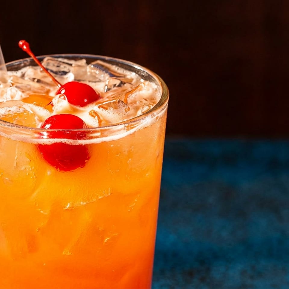 Close-up of a vibrant orange cocktail with ice, two maraschino cherries, and a straw.