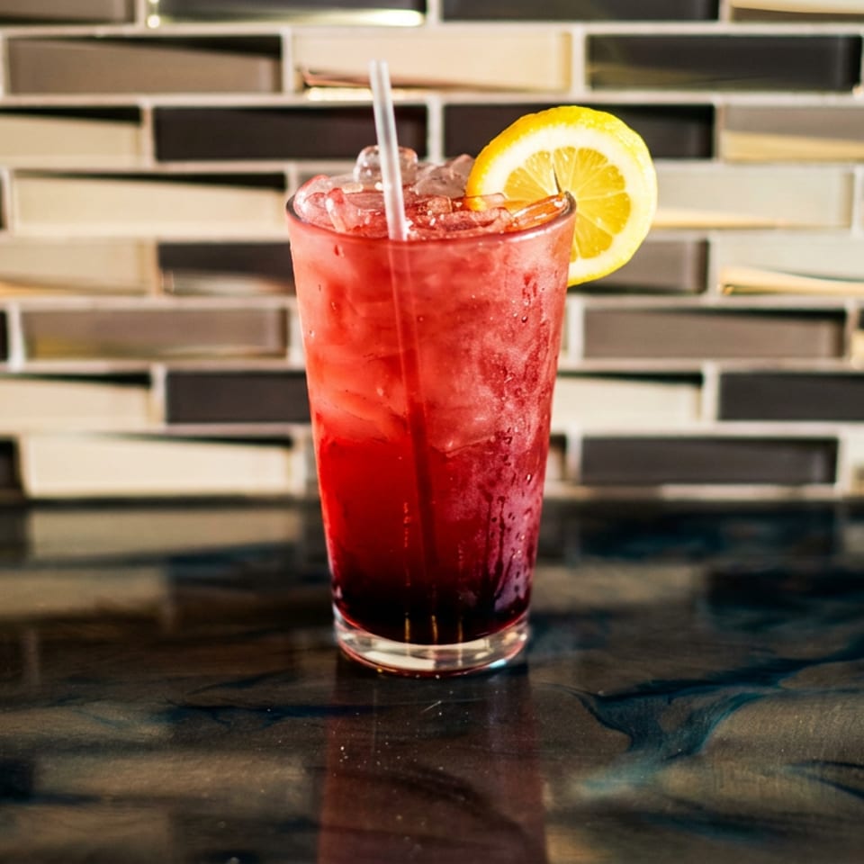 A vibrant red cocktail with ice, a straw, and a lemon slice sits on a dark, reflective counter.