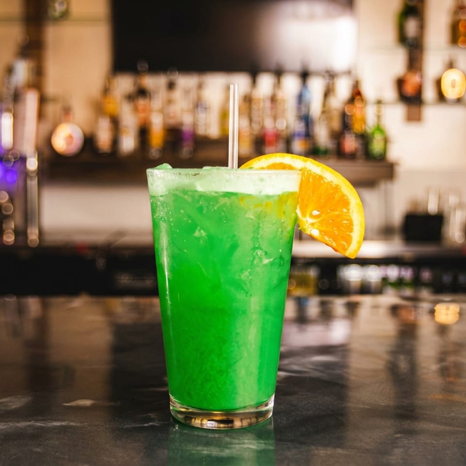 A vibrant green cocktail with an orange slice and straw sits on a dark bar counter.