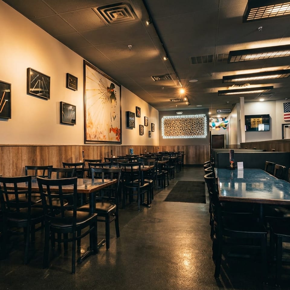 Empty restaurant dining room with dark tables and chairs, wood paneling, and wall art.