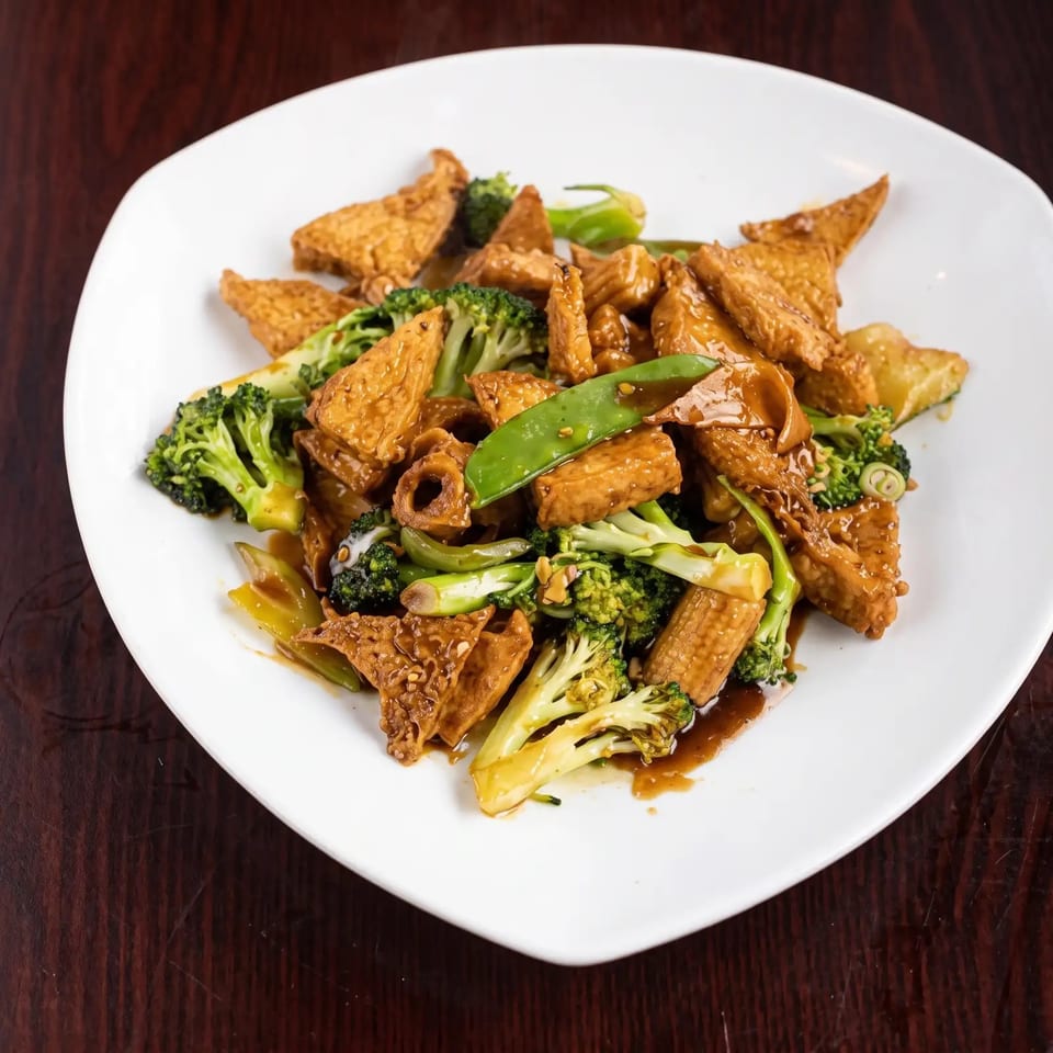 Stir-fried tofu, broccoli, baby corn, and snow peas in a savory sauce on a white plate.