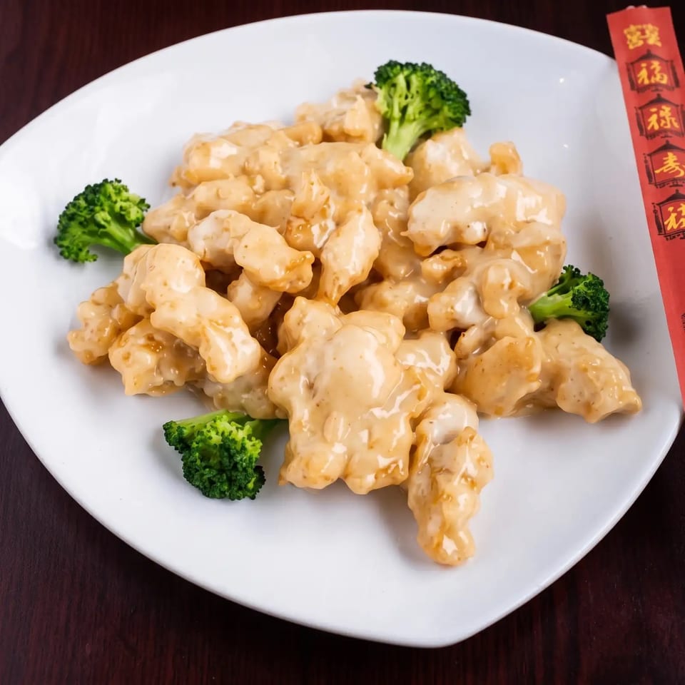 Creamy orange chicken with broccoli florets on a white plate, with red chopsticks.
